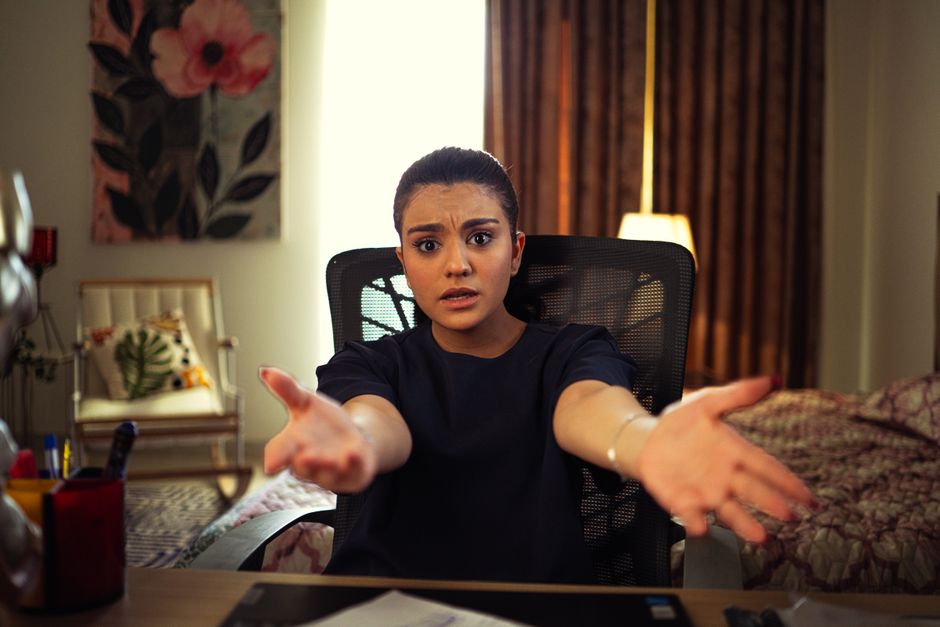 A young woman seated indoors, extending arms in an expression of frustration.