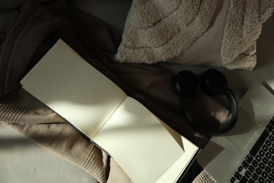A warm, minimalist flat lay of an open notebook, headphones, and laptop on a bed with soft lighting.