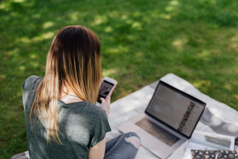 From above back view of skinny long haired teenage girl in casual clothes browsing social media on smartphone while spending time in sunny park during summer vacation