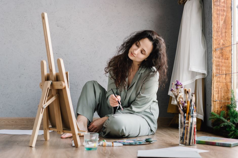 A woman in comfortable attire paints leisurely by a window with art supplies around her.