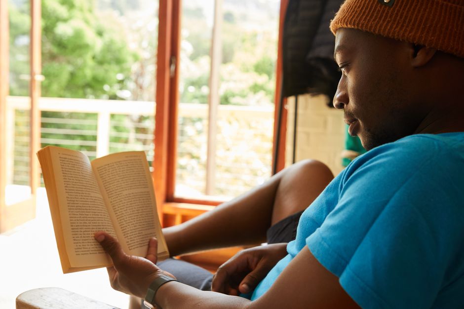 Side view of crop black male in trendy hat resting on comfortable couch while reading interesting book in apartment