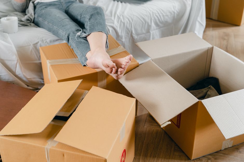 Crop faceless barefoot female in jeans sitting with crossed legs on cozy couch covered with crumpled bed sheet near cup of hot drink and pile of cardboard boxes on floor