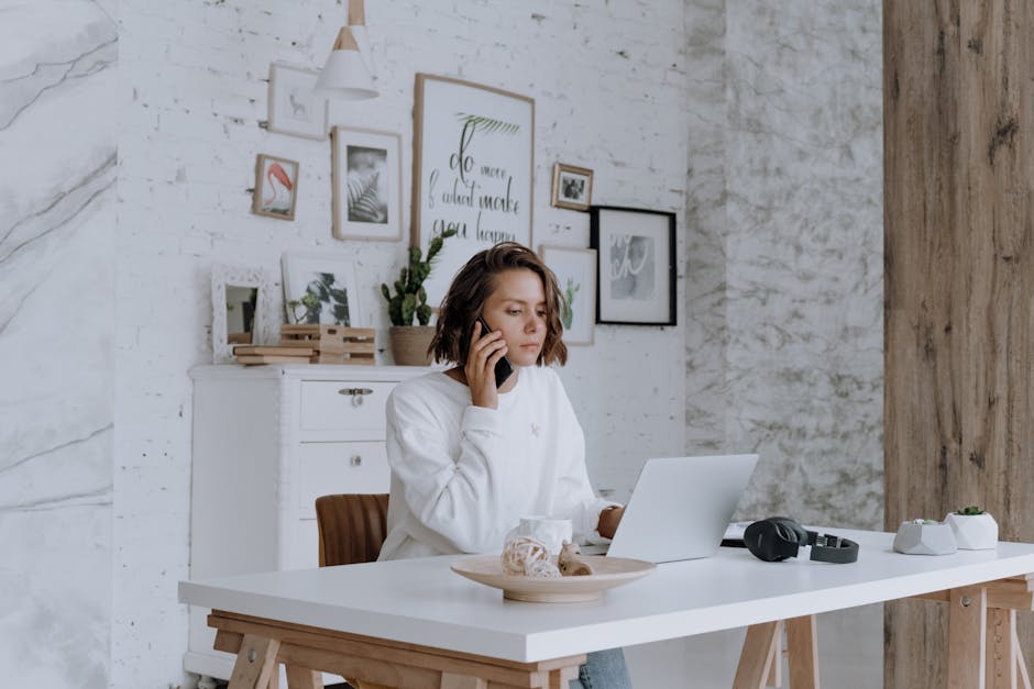 Woman talking on phone while using laptop in bright, modern home office with decor.