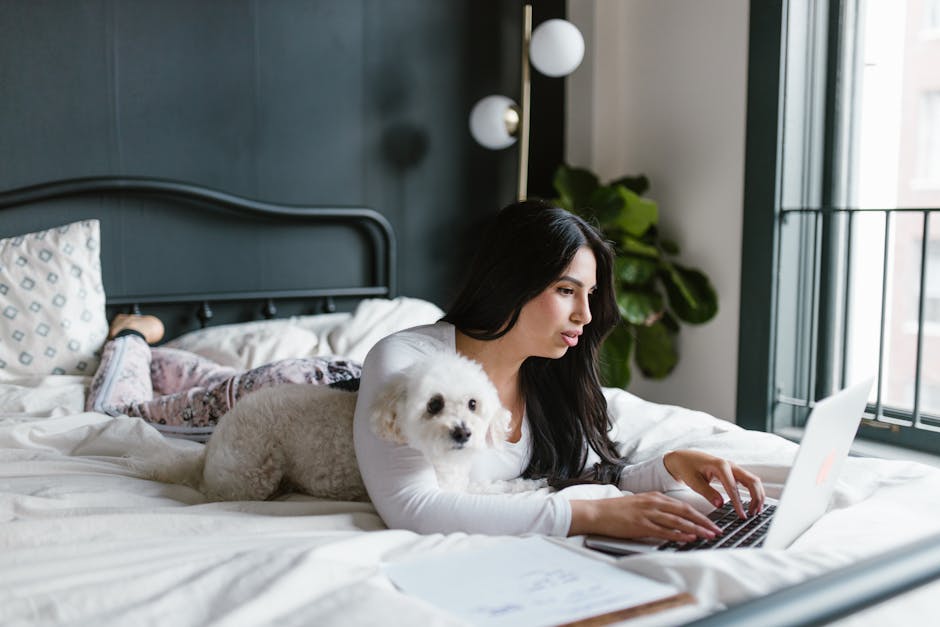 Woman working remotely on laptop with her dog, lying on a bed in a cozy bedroom setting.