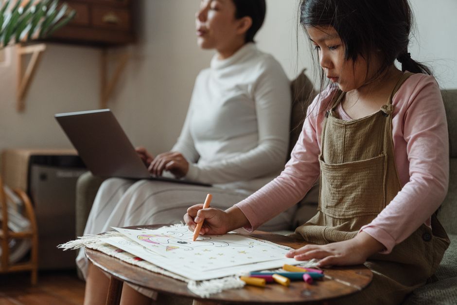 Crop cute concentrated Asian girl sitting at table and coloring picture with crayons near focused mother working on netbook on sofa in light living room