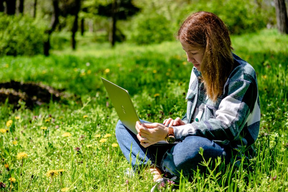 A woman sitting on grass using a laptop, enjoying remote work in a sunny park setting.